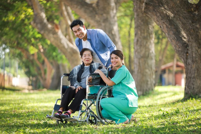 車椅子に乗った高齢の女性と車椅子を押す若い男性、しゃがんで並ぶ介護施設の若い女性