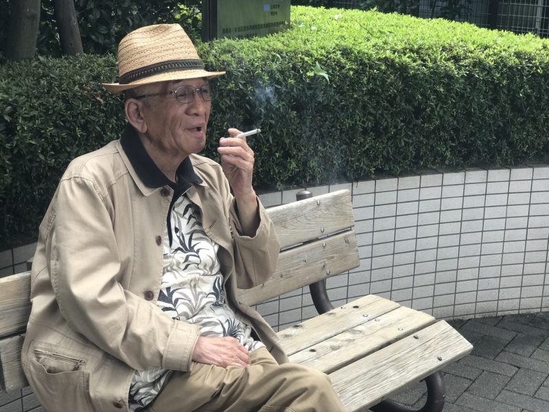 ベンチに腰掛けタバコを吸う矢崎氏の画像。タバコと帽子は矢崎氏にとって欠かせないもの