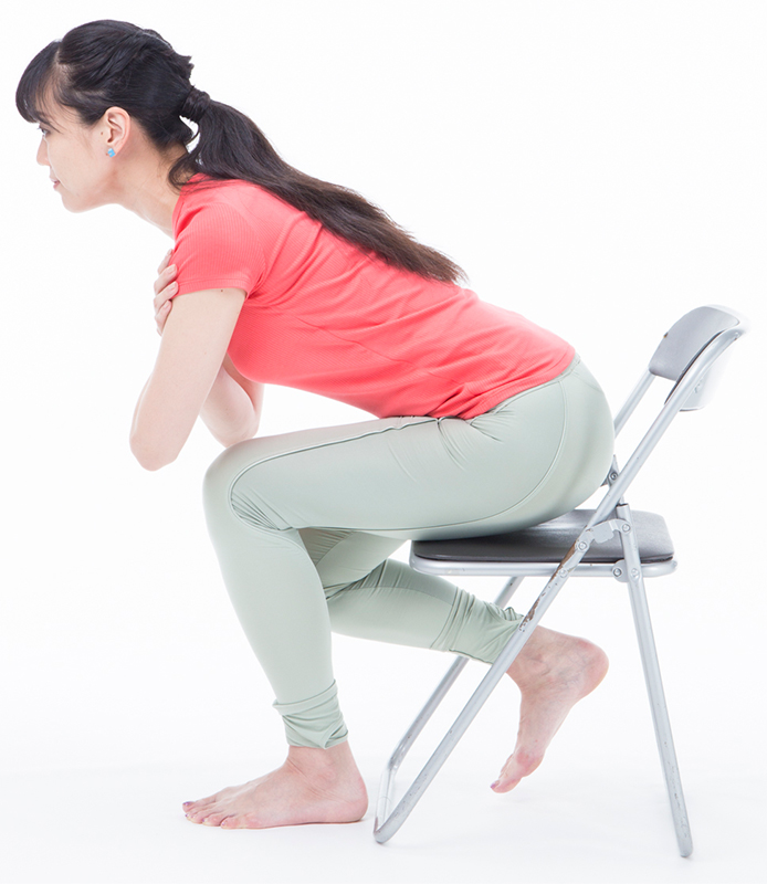 椅子に腰かけ、手を胸の前で組む。しっかりと前傾し、立つ脚に全体重をのせる。