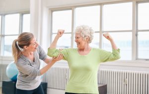Portrait of female trainer looking at elderly woman flexing her bicep at the rehabilitation center. Happy about her recovery at rehab.