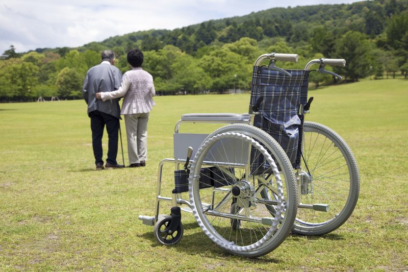 杖を突いて歩く高齢男性と支える高齢女性、その手前に車いすが置いてある