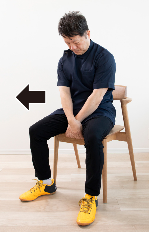 椅子に浅く座り、太ももの間で両手をクロスさせて椅子の前部分をつかむ男性の写真