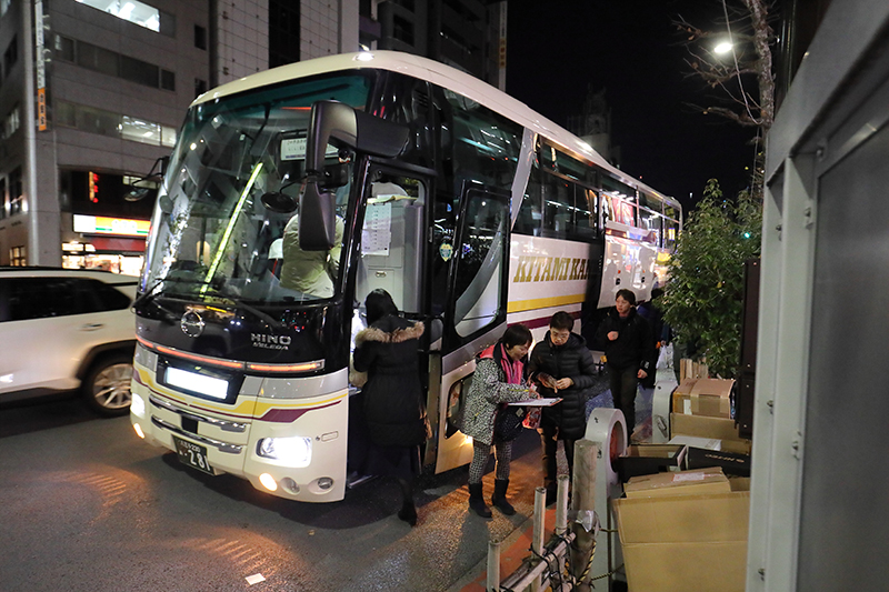 夜7時、大型バスとその入り口でツアーの受付する添乗員とお客