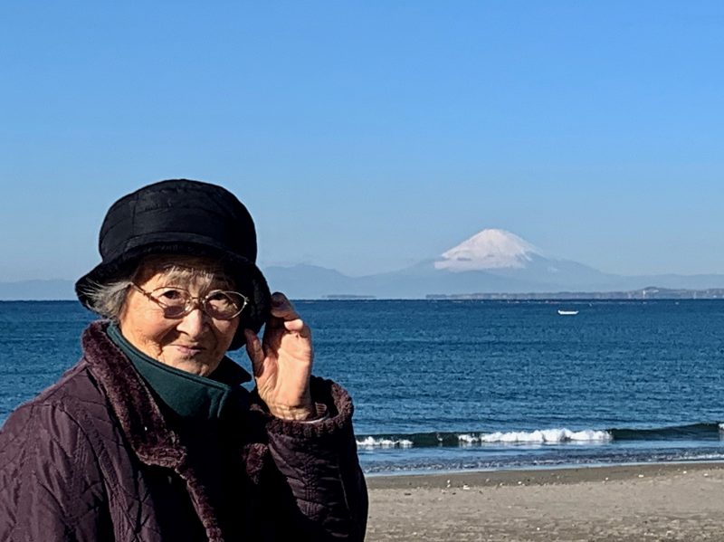 富士山が遠くに見える内房の海岸と帽子をかぶった高齢の女性