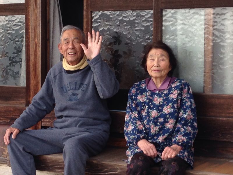 縁側に座って手を挙げる高齢の男性とその妻
