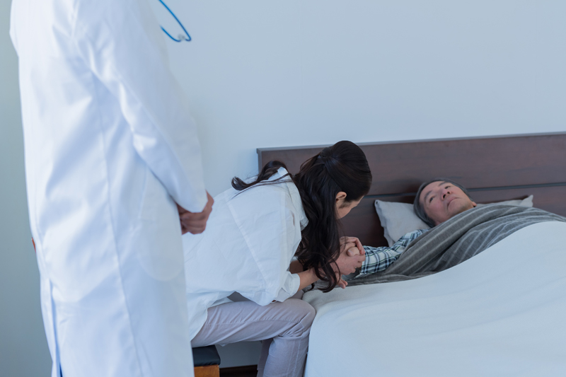 ベッドに横たわる男性の手を取る女性と、傍らに立つ医師の写真