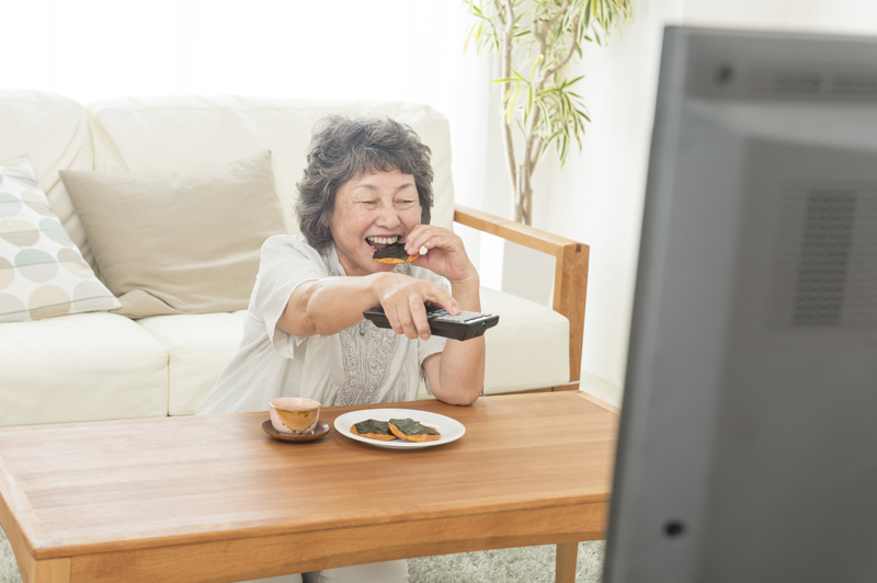 おせんべいを食べながら、笑ってテレビを見るシニア女性
