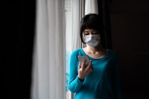 Indoor portrait of Asian woman wearing surgical mask operating smartphone at the window